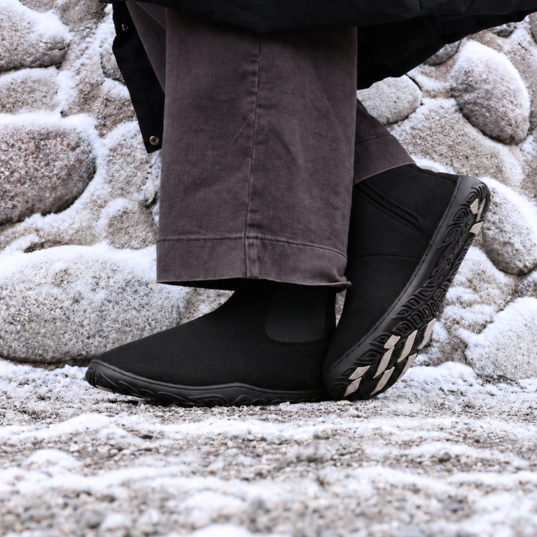 Vaistoa Sulo 2 barfota chelsea-boots för vinter – livsstilsbild: paret på en snöig klippa med mörkbruna byxor, ena skon lutad så att greppdynorna på yttersulan syns, en stenmur i bakgrunden