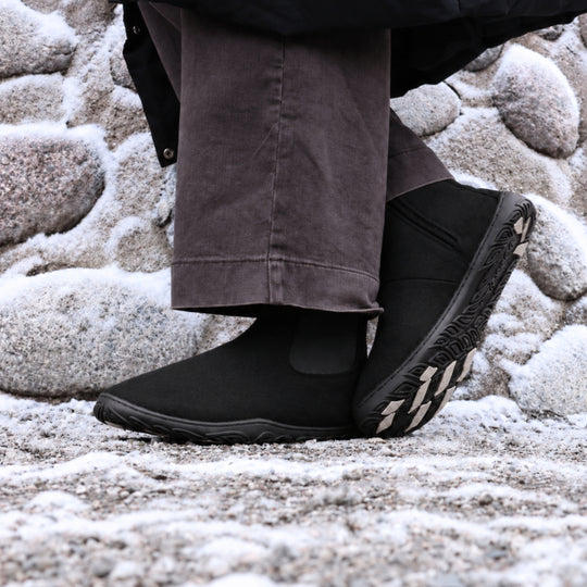 Vaistoa Sulo 2 barfota chelsea-boots för vinter – livsstilsbild: paret på en snöig klippa med mörkbruna byxor, ena skon lutad så att greppdynorna på yttersulan syns, en stenmur i bakgrunden