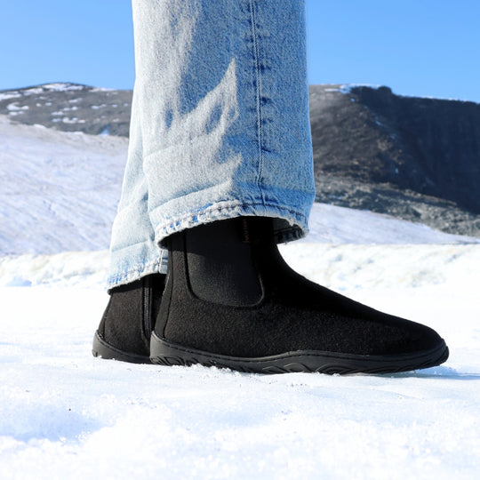 Vaistoa Sulo 2 barfota chelsea-boots för vinter – livsstilsbild: skon från sidan på snön med blå jeans, den elastiska resåren syns, ett fjäll och blå himmel i bakgrunden