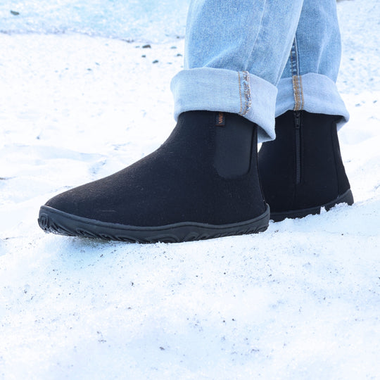 Velvet black barefoot winter felt Chelsea boots worn with blue jeans in a snowy winter setting, shown from a front angle to highlight their warm felt texture, flexible sole, and natural barefoot design.