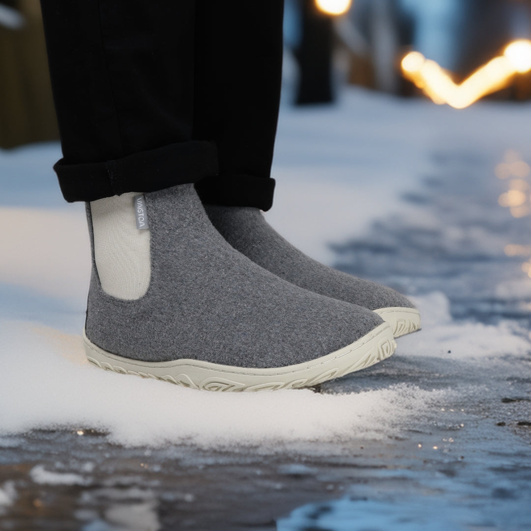 Käyttökuva, jossa henkilö seisoo helmenharmaat Vaistoa Sulo -huopanilkkurit jalassa lumisella kadulla iltavalojen loisteessa. Kuvakulma on etuviistosta ja korostaa kengän luonnollista muotoilua ja pohjan pitoa märällä alustalla.