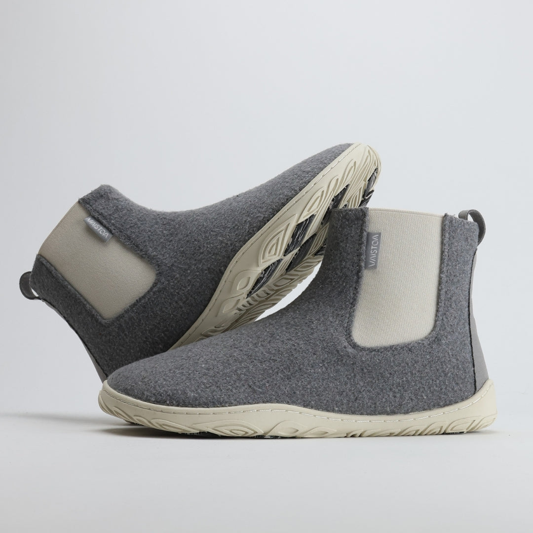 Kaksi helmenharmaata Vaistoa Sulo -huopanilkkuria, joista toinen nojaa toisen varteen. Kuvassa näkyy kengän sivuprofiili ja pohjan kitkapalat, jotka on suunniteltu tarjoamaan varmuutta liukkaalla alustalla.