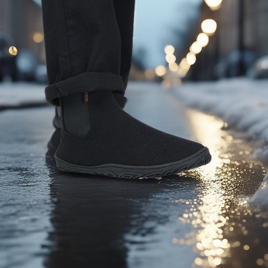 Kuva henkilöstä, jolla on jalassaan samettisen mustat Vaistoa Sulo -huopanilkkurit. Kuvattu sivulta iltahämärässä, katuvalojen loisteessa, korostaen pohjan pitoa ja luonnollista askellusta märällä pinnalla.