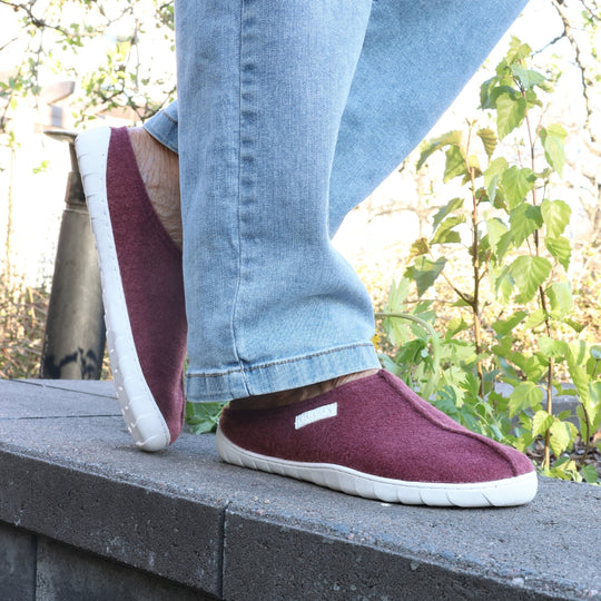 Vaistoa Toive felt barefoot slippers – burgundy felt slippers worn outdoors on a stone edge, light blue jeans and green plants in sunny spring or autumn weather