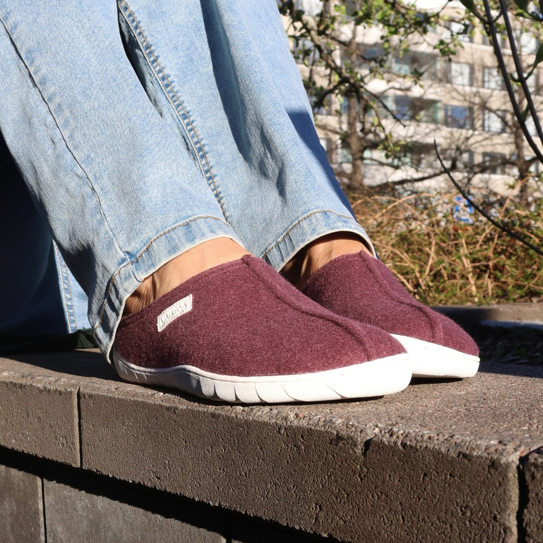 Vaistoa Toive felt barefoot slippers – burgundy felt slippers worn outdoors on a stone edge, light jeans and a sunny yard with green plants in the background