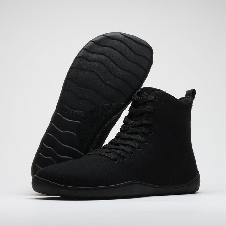 Vaistoa Tunto 2 felt barefoot sneakers – studio shot of the pair on a light background, one shoe leaning on the other with its outsole facing the camera, showing the wavy tread pattern and embossed VAISTOA lettering, the front shoe upright with its lacing and high collar visible, black colour