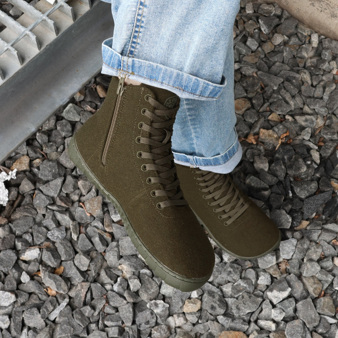 Vaistoa Tunto 2 felt barefoot sneakers – on-foot, blue jeans, seated on a metal grating above gravel, side zipper visible, olive colour
