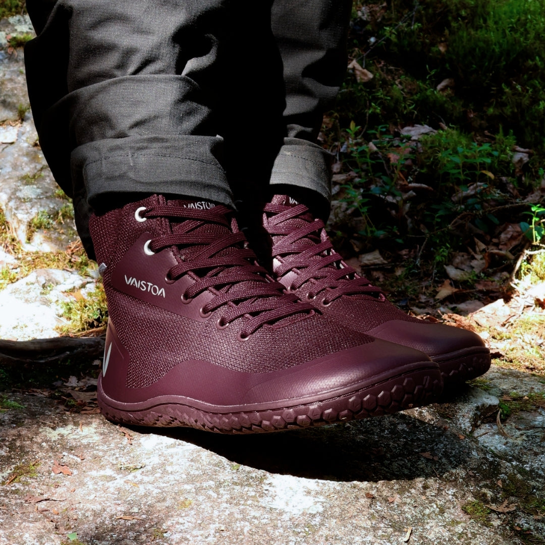 Vaistoa Tuohi 2 barefoot light hiking shoes burgundy – close-up of the shoes being worn, the wearer standing on a rock covered in pale crustose lichen, with moss around it