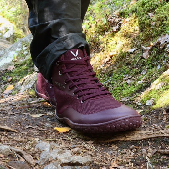 Vaistoa Tuohi 2 barefoot light hiking shoes burgundy – close-up of the shoes being worn, the wearer standing on a damp forest trail with moss, stones and a yellow birch leaf on the ground