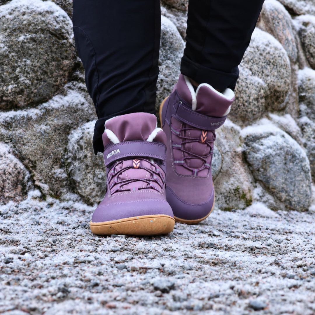 Vaistoa Ulappa 2 kids waterproof barefoot sneakers – lifestyle photo: a step on frosty ground from the front with black trousers, the hook-and-loop strap, quick lacing and faux fur lining visible, rocks in the background