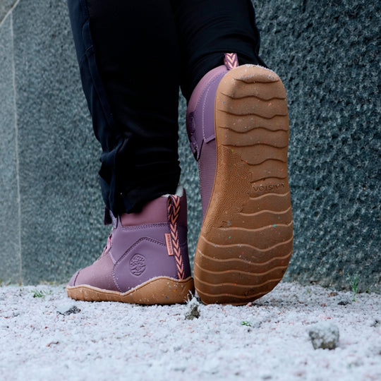 Vaistoa Ulappa 2 kids waterproof barefoot sneakers – lifestyle photo: a step on frosty ground with black trousers, the wave-patterned outsole of the back shoe with its colourful recycled-material specks visible, a stone wall in the background