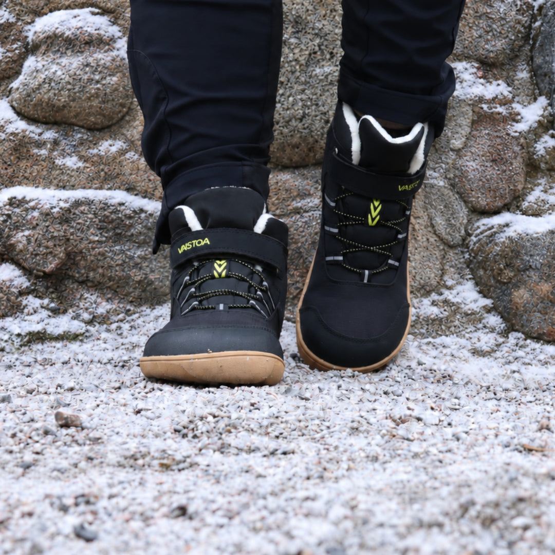 Vaistoa Ulappa 2 kids waterproof barefoot sneakers – lifestyle photo: a step on frosty ground from the front with black trousers, the hook-and-loop strap, quick lacing and faux fur lining visible, rocks in the background