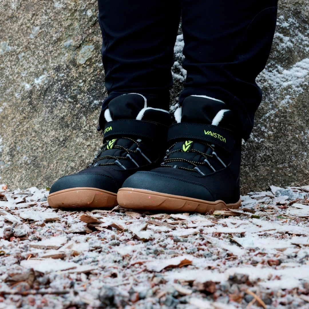 Vaistoa Ulappa 2 kids waterproof barefoot sneakers – lifestyle photo: pair from the front on a frosty gravel yard in front of a rock face with black trousers, the Vaistoa logo on the hook-and-loop strap, quick lacing and faux fur lining visible