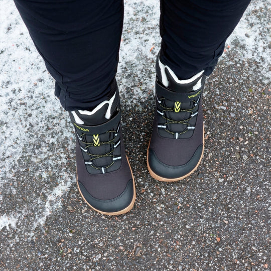 Vaistoa Ulappa 2 kids waterproof barefoot sneakers – lifestyle photo: pair from above standing on frosty asphalt with black trousers, the hook-and-loop strap, quick lacing and faux fur lining visible
