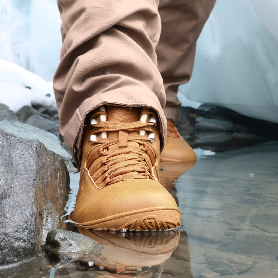 Vaistoa Uoma 4 winter barefoot trekking boots – lifestyle photo: a step on a wet rock by the water from a front angle with beige trousers, lacing and faux fur lining visible, ice in the background