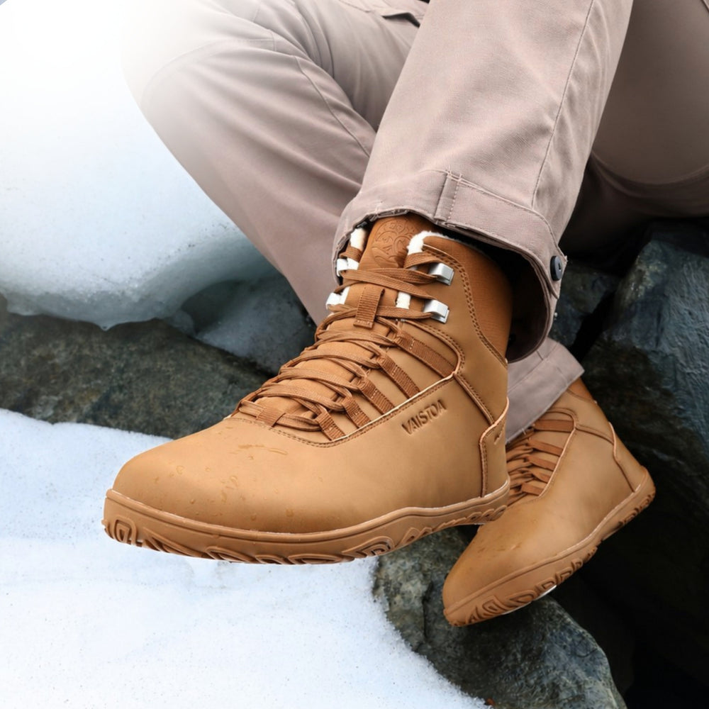 Vaistoa Uoma 4 winter barefoot trekking boots – lifestyle photo: pair worn on snowy rocks from a front angle with beige trousers, the wearer seated, lacing and faux fur lining visible