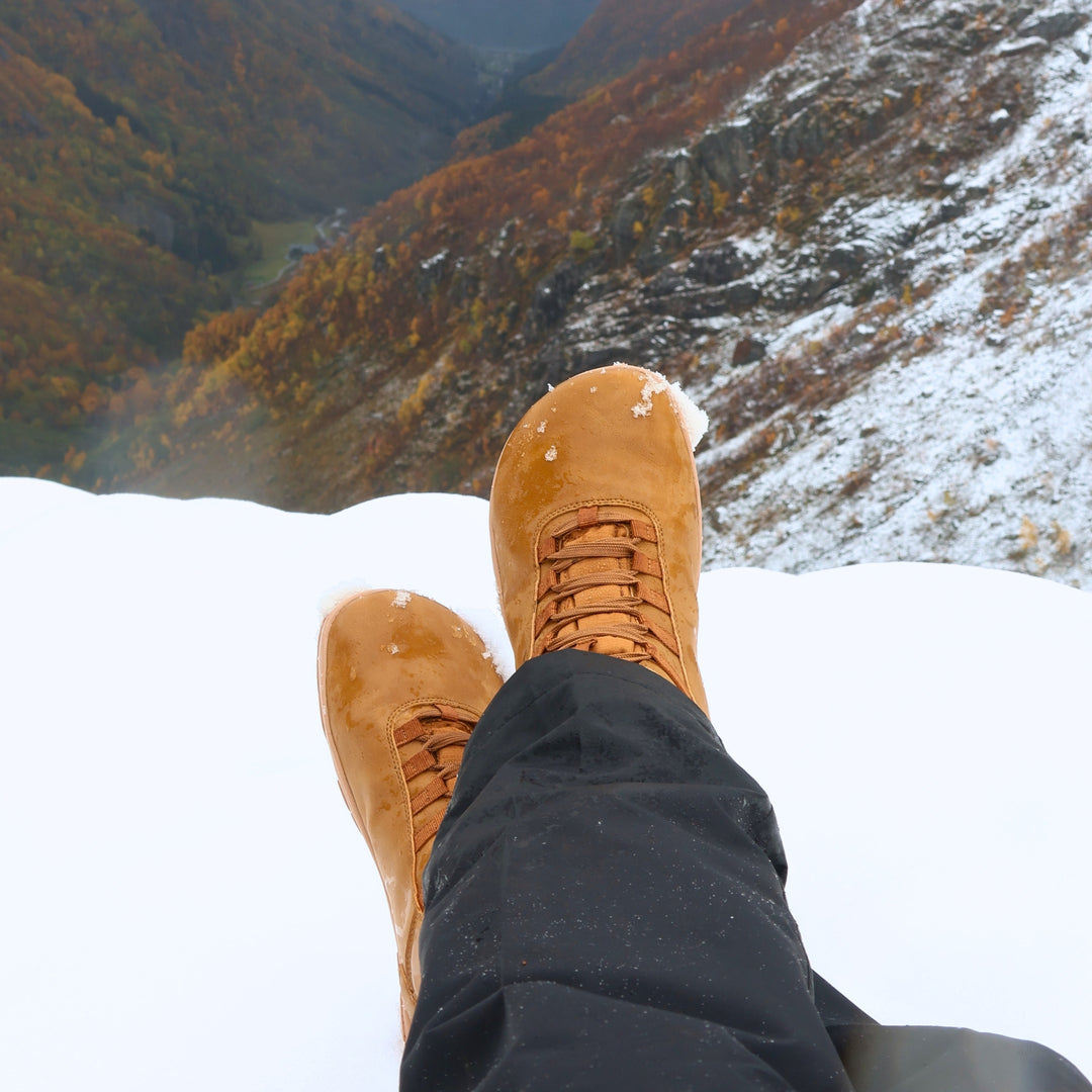 Vaistoa Uoma 4 winter barefoot trekking boots – lifestyle photo: feet resting on snow with black trousers, a mountain valley in autumn colours in the background