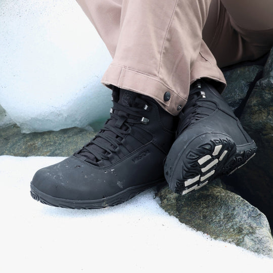Vaistoa Uoma 4 winter barefoot trekking boots – lifestyle photo: pair worn on a snowy rock with beige trousers, the seated wearer crosses one foot so the outsole with light grip pads is visible, ice in the background