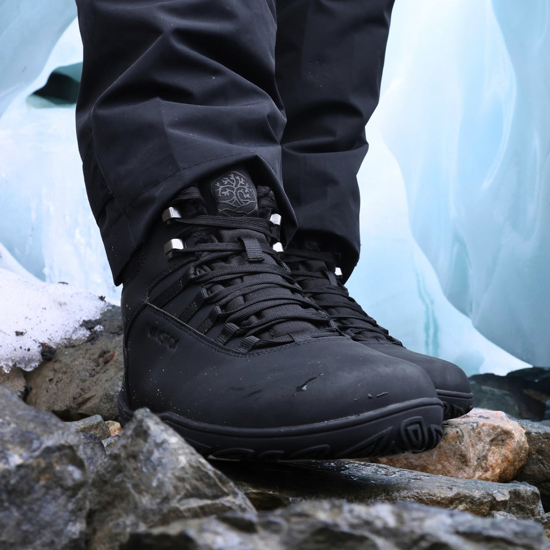 Vaistoa Uoma 4 winter barefoot trekking boots – lifestyle photo: pair worn on rocks shown close up from a front angle with black trousers, lacing and metal lace hooks visible, an ice wall in the background
