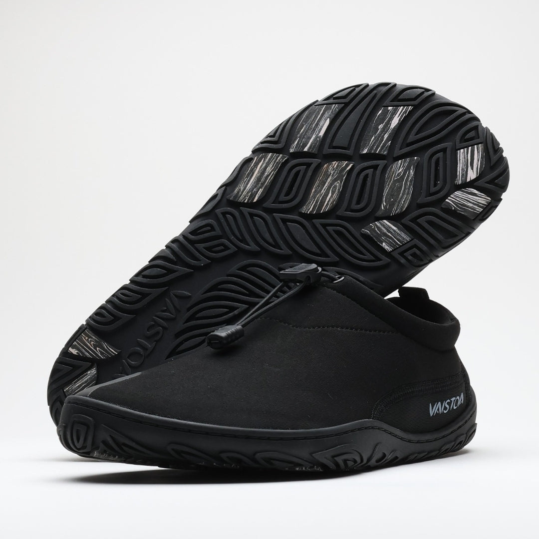 Black Vaistoa Vire barefoot shoe shown from the side. Another shoe is placed upside down in the background, revealing the organic outsole pattern and light-colored grip pads. Low-profile, flexible construction with a quick-lace system. Design from Finland label in the top right corner.