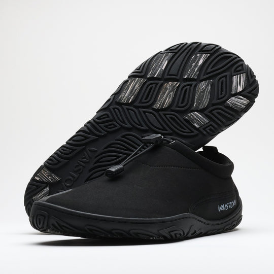 Black Vaistoa Vire barefoot shoe shown from the side. Another shoe is placed upside down in the background, revealing the organic outsole pattern and light-colored grip pads. Low-profile, flexible construction with a quick-lace system. Design from Finland label in the top right corner.