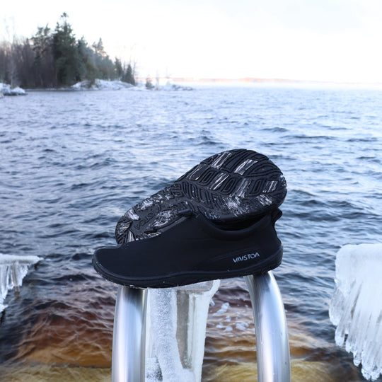 Black Vaistoa Vire barefoot shoe placed on the railing of an ice swimming pier next to icy water. The outsole of one shoe is visible, with the outsole pattern and grip pads clearly standing out in ice swimming conditions.