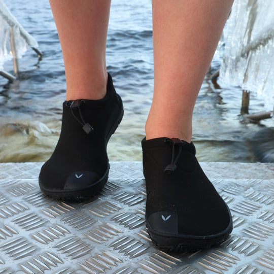Black Vaistoa Vire barefoot shoes worn on feet, shown from the front on an ice swimming pier. Both shoes are visible, highlighting the flexible neoprene upper and quick-lace system. Ice hole and cold water in the background.