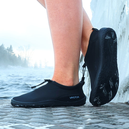 Black Vaistoa Vire barefoot shoes worn on feet, shown from the side on an ice swimming pier. The outsole of one shoe is visible, clearly showing the organic outsole pattern and light-colored grip pads. Icy water in the background.