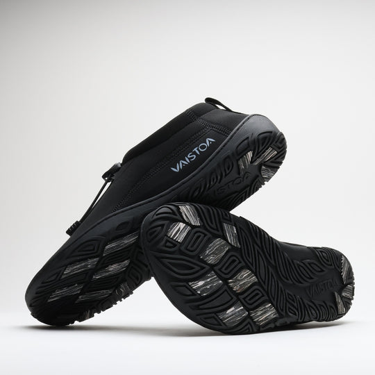 Black Vaistoa Vire barefoot shoes shown in a studio setting with both outsoles fully visible. The outsole pattern and light-colored grip pads stand out clearly against the light background.